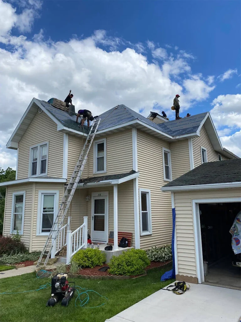 Workers installing a new roof on a beige house, with a blue sky and fluffy clouds above, and a ladder leaning against the side.
