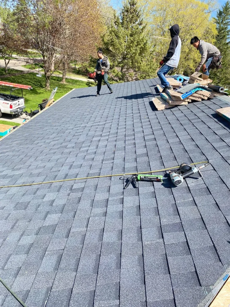 Three roofers working on a blue shingle roof, with tools scattered around, under a clear sky and surrounded by trees.