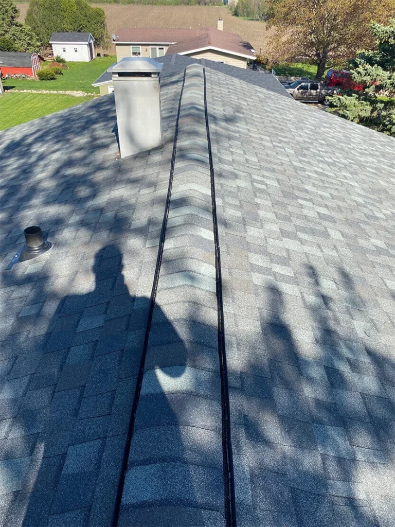 A rooftop view showcasing gray shingles, a chimney, and shadows, with green fields and structures in the background.