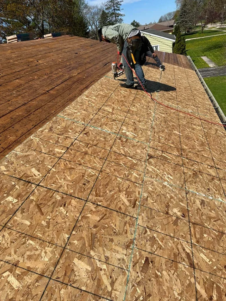 Two workers install plywood sheets on a sloped roof, using pneumatic tools under a clear blue sky.