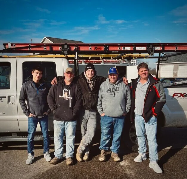 Five men stand side by side in front of a white work truck with a red ladder on top, posing for a group photo on a sunny day.