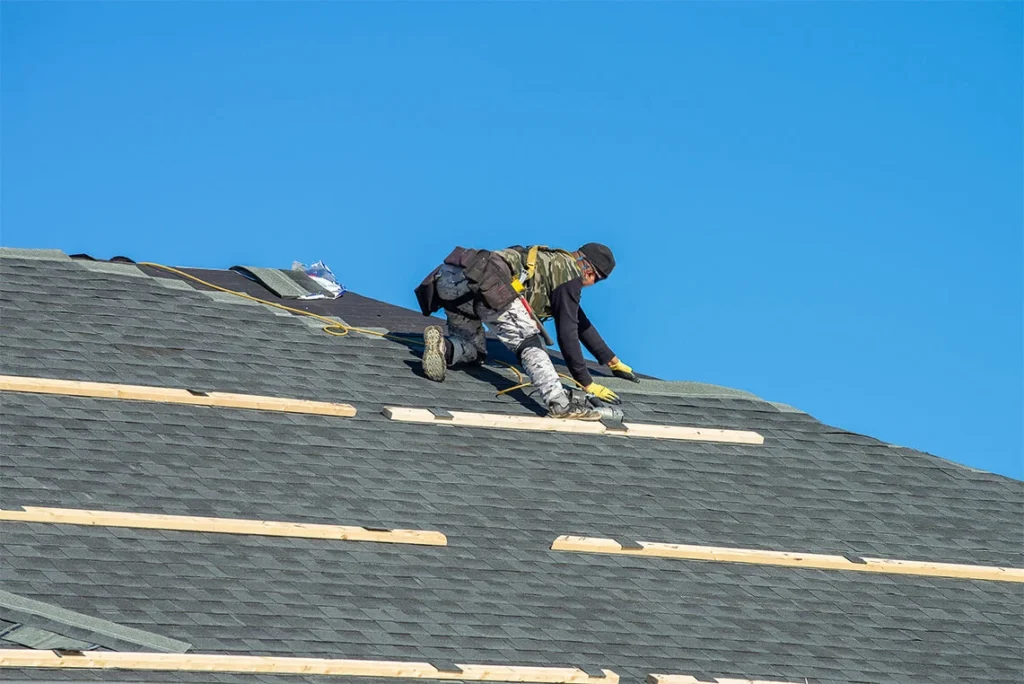 A worker in safety gear installs shingles and wooden battens on a sloped roof under a clear blue sky.