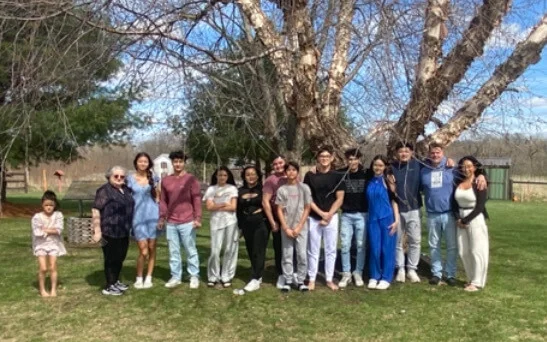 A diverse group of 15 people, including children and adults, pose together outdoors by a large tree on a sunny day.