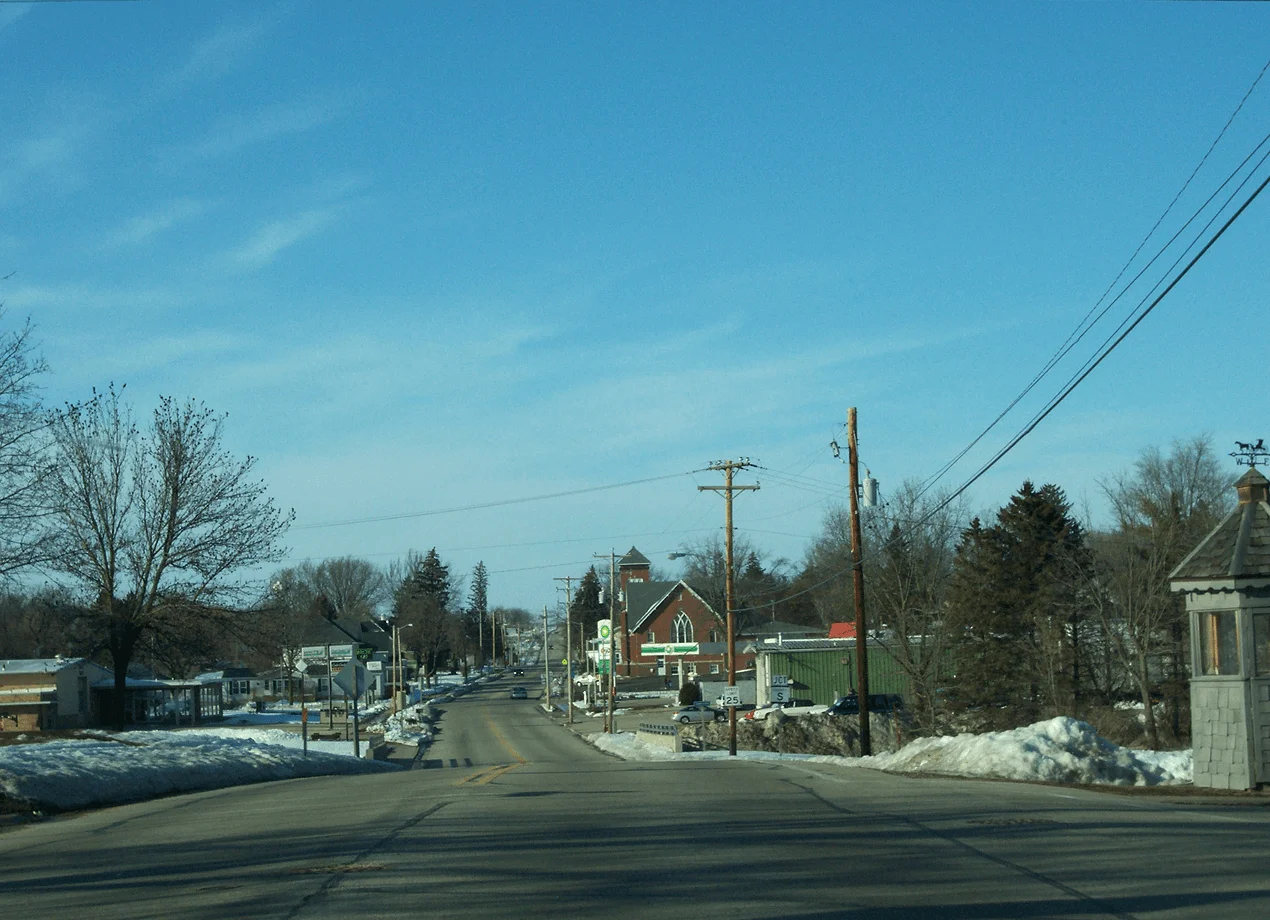 A quiet winter street scene with snowbanks, power lines, and small buildings under a clear blue sky. A church steeple is visible in the distance.