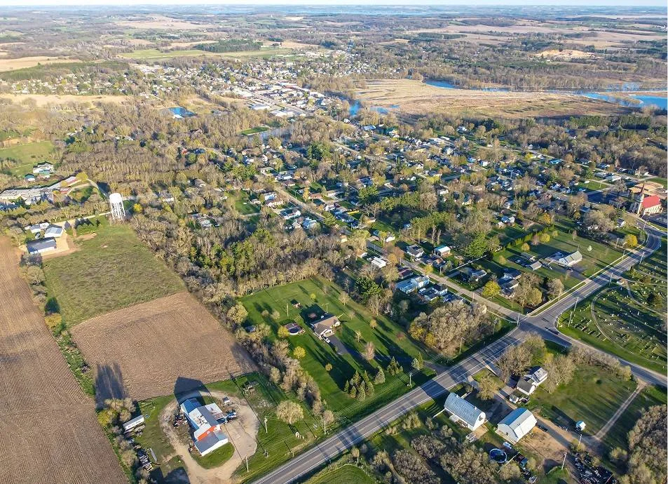 Aerial view of a small rural town with houses, a water tower, farmland, and a winding road surrounded by trees and open fields.