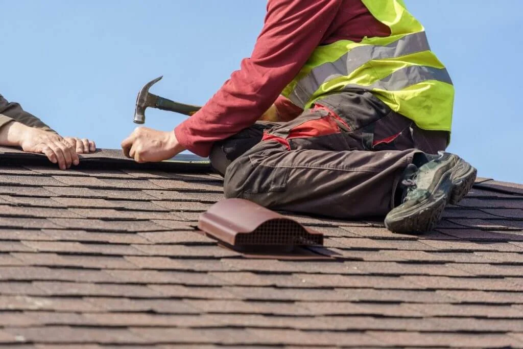 A man sitting on a roof with a hammer in his hand, surrounded by a clear blue sky and the structure of the building around him. He is wearing casual clothing.