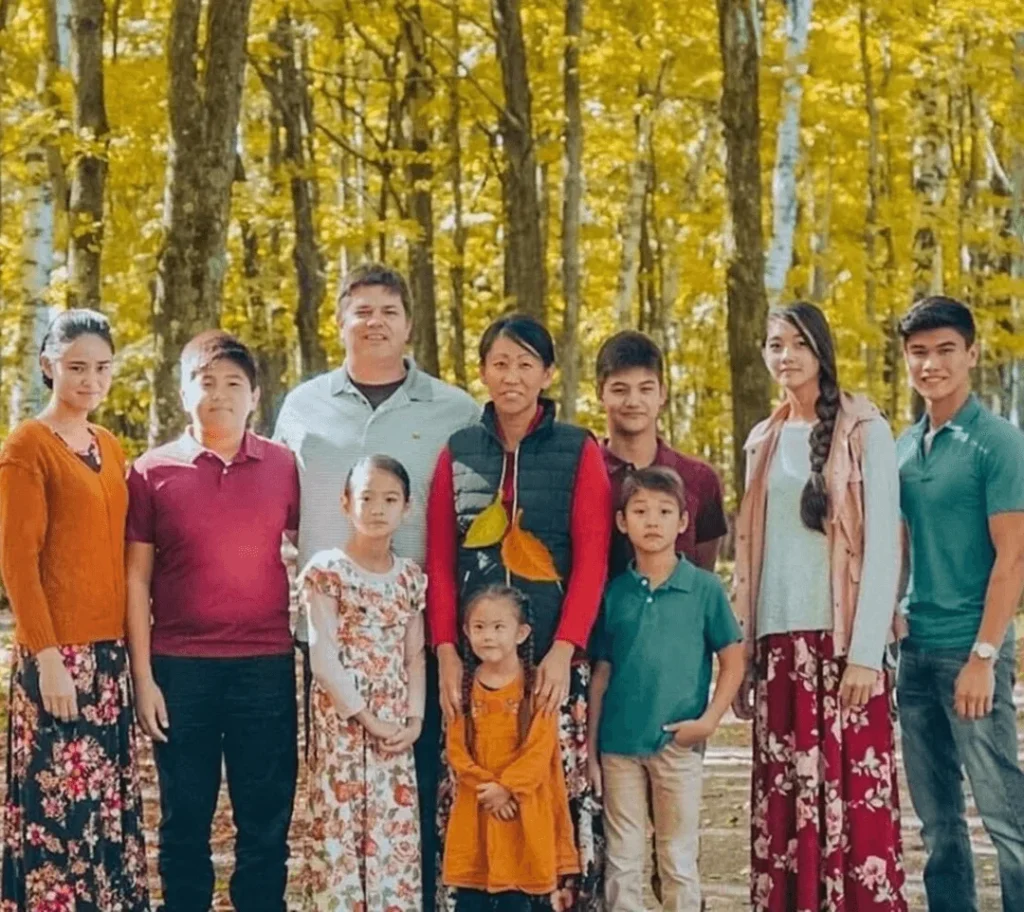 A group of people standing together outdoors, smiling for a photo. They are dressed in various clothing styles, and there is a tree in the background. The group includes boys and girls of different ages, with some wearing dresses.