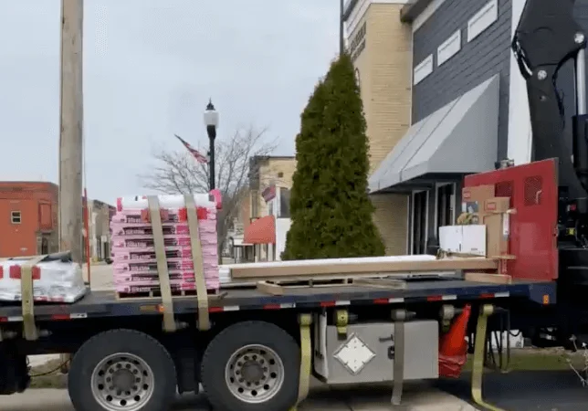 A flatbed truck parked outdoors, loaded with boxes, on a road under a clear sky. The truck features large wheels and is designed for transport.