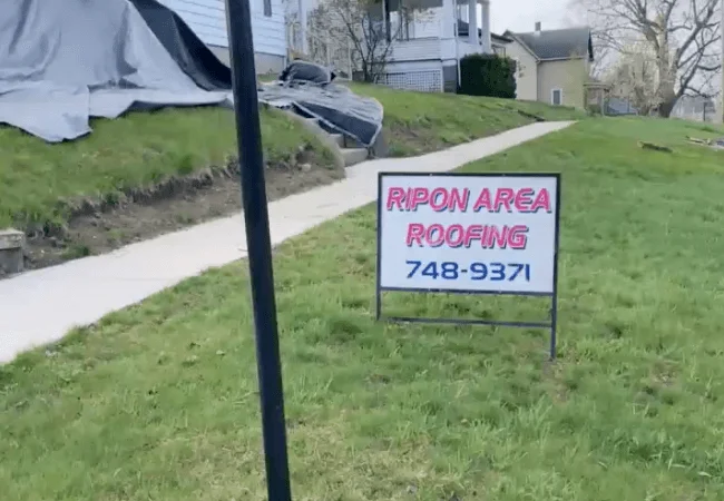 A sign in the grass displaying "RIPON AREA ROOFING 748-9371". The sign is surrounded by green grass, with trees visible in the background.