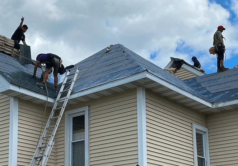 Five roofers work on a residential home, installing shingles under a partly cloudy sky, with ladders and materials visible.