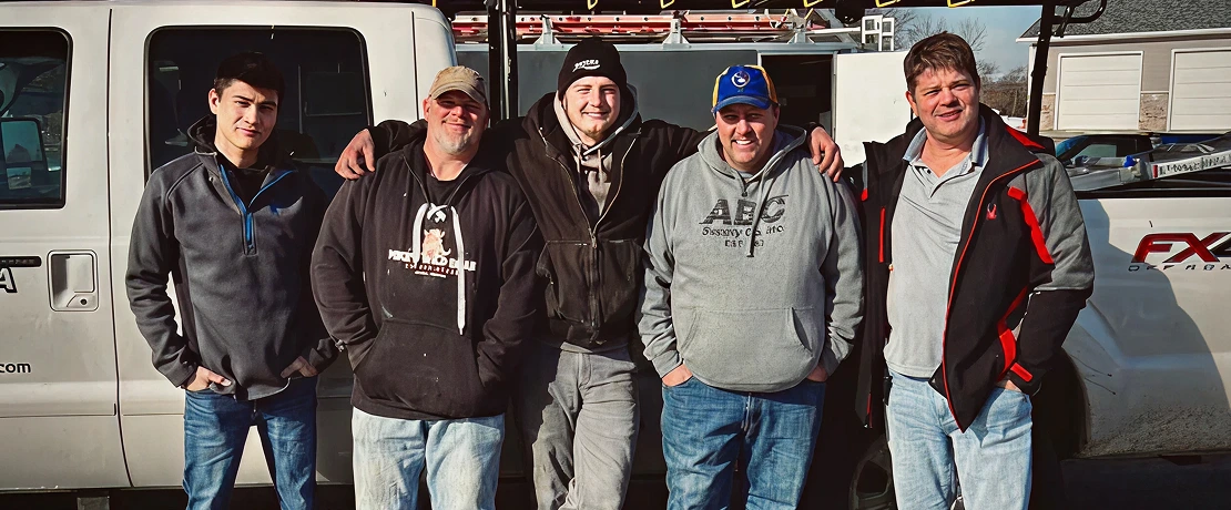 Five smiling roofing contractors standing in front of a work truck, showcasing a friendly and professional team ready to serve