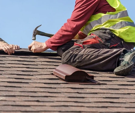Roofer in high-visibility vest using a hammer while installing ridge cap on a brown shingle roof.