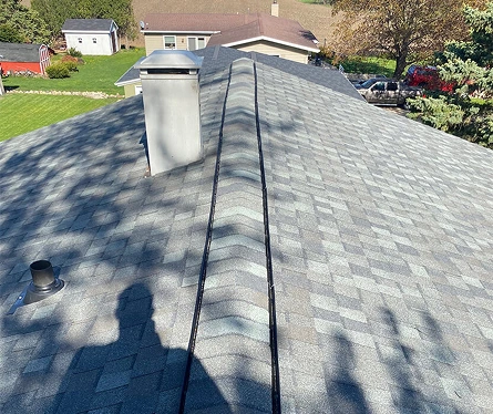 View of a newly completed gray shingle roof with ridge vent, chimney, and surrounding suburban landscape.